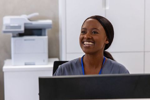 Smiling receptionist greeting patients at clinic reception desk wearing scrub and lanyard
