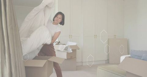 Korean woman packing duvet into box during move in neutral bedroom with wardrobe