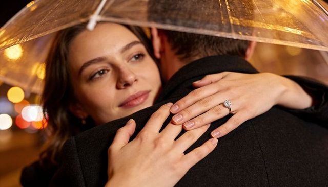 Engaged couple embracing under clear umbrella showing solitaire diamond ring on rainy city street