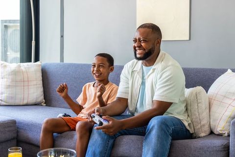 African american father and son playing video games on gray sofa, family bonding at home