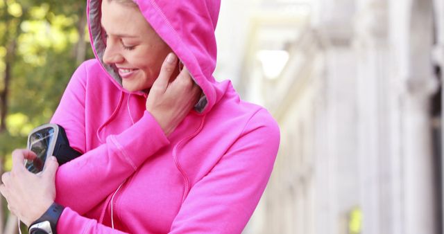 A young Caucasian woman in a pink hoodie is setting up her music player before a run, with copy space. Her focus on the device underscores the importance of a motivating playlist for an enjoyable workout.