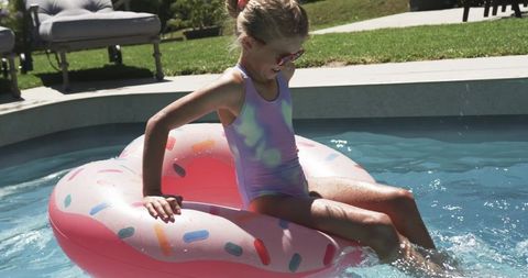 Young girl sitting on pink donut float splashing in backyard pool wearing pastel swimsuit