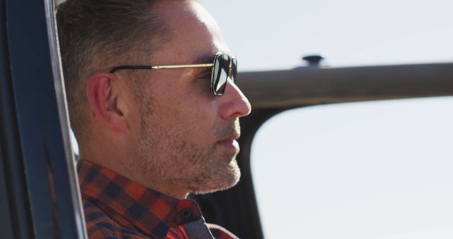 Man with sunglasses in car, relaxing and enjoying daytime. Ideal for themes related to summer road trips, relaxation, casual fashion, and outdoor activities.