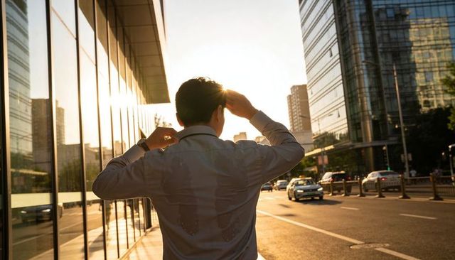 Businessman adjusting loosened tie on urban sidewalk at golden hour with sweat stains