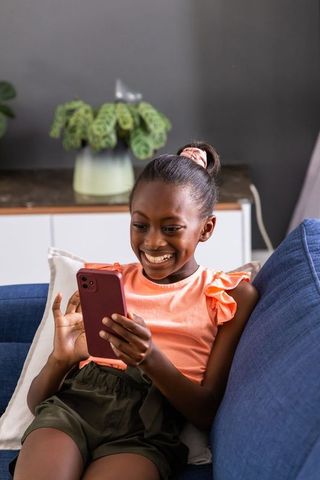 Smiling african preteen girl sitting on blue couch holding pink smartphone and using app