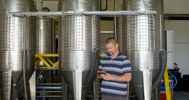 Man using tablet in modern brewery with large metal tanks. Ideal for industrial processes, craft beer, brewery equipment, and production technology topics.