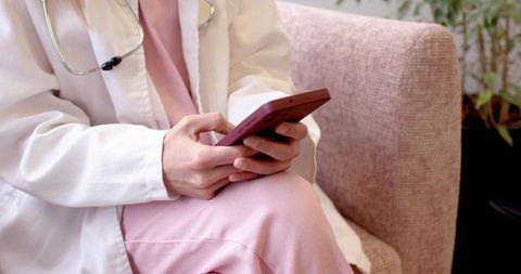 Female clinician using smartphone while seated in pastel clinic lounge wearing white coat