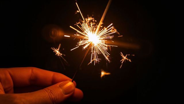 Closeup hand holding sparkler at night with bright golden sparks and lens flare