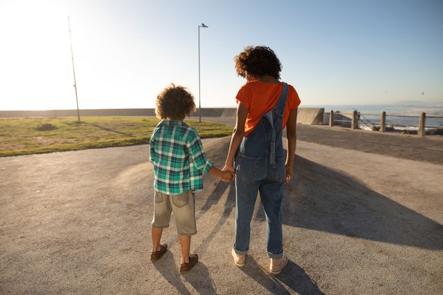 Rear view of a biracial woman wearing denima dungarees and her son enjoying time together by the sea, holding hands and smiling at each other at a playground on a sunny day