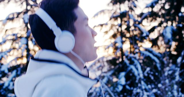 A young man is listening to music with his headphones while standing in a snowy winter forest. He is wearing a white jacket and appears calm and contemplative. This image is ideal for promoting winter activities, music streaming services, outdoor recreation, or portraying serenity and relaxation in nature.