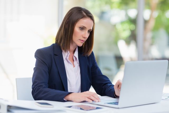 Businesswoman in a suit working on a laptop in a modern office environment. Ideal for use in business, technology, and professional career-related content. Can be used for articles, presentations, and websites focusing on corporate life, productivity, and professional development.