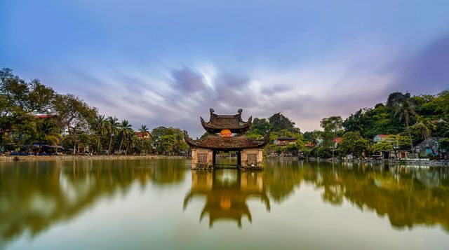 A stunning view of an ancient Vietnamese temple at sunset reflecting on a serene lake. The vibrant colors of the sky add to the picturesque scenery. Ideal for use in travel brochures, cultural documentaries, tourism websites, and wallpaper backgrounds.