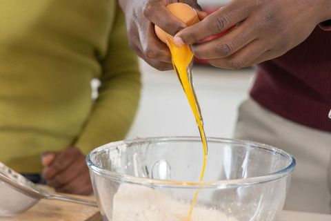African american friends cracking egg into bowl while baking together in home kitchen