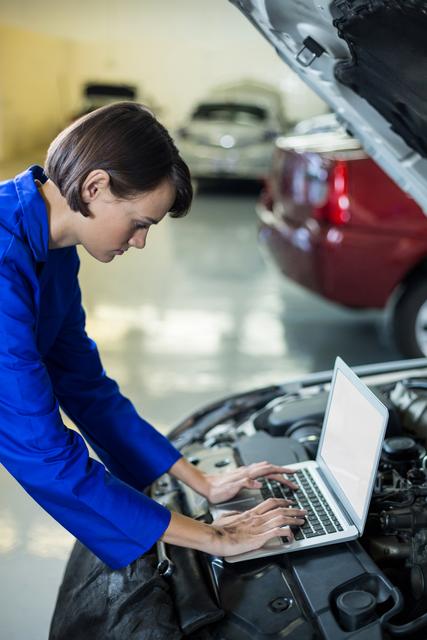 Female mechanic using laptop to diagnose car engine issues in a repair garage. Useful for content related to auto repair services, the role of women in technology and mechanics, automotive diagnostics, and professional service promotion.
