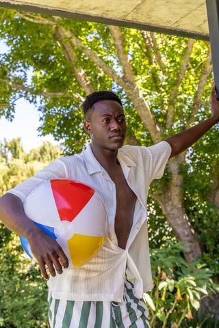 African american man holding colorful beach ball, wearing open shirt and striped trunks