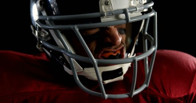 A close-up of a determined American football player in a helmet looking focused. The player is wearing a red jersey. This image is perfect for illustrating themes such as sportsmanship, focus, and intensity in competitive sports. It can be used in promotional material for sports events, articles on football techniques, or motivational content emphasizing determination.