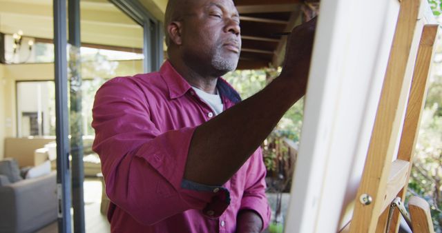 Happy african american senior man standing on balcony painting picture on canvas. retirement lifestyle, spending time alone at home.