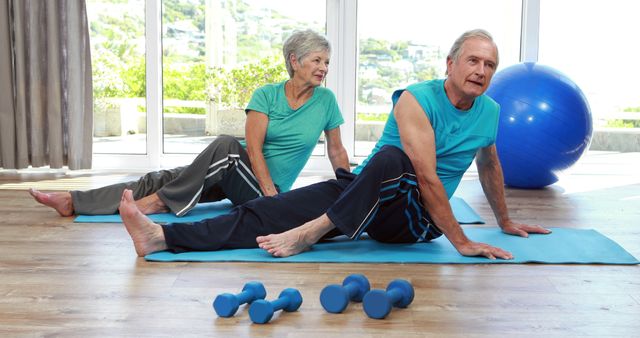 Senior couple is stretching and exercising on yoga mats in a bright room. They have dumbbells on the floor, showing their commitment to maintaining an active and healthy lifestyle. Perfect for promoting senior fitness, healthy aging, wellness programs, and active retirement living.