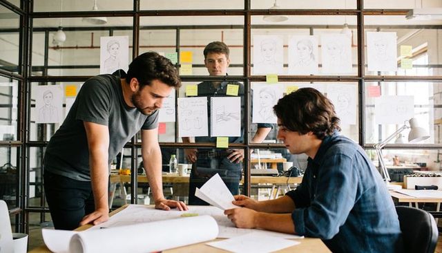 Colleagues collaborating over blueprints in creative studio with sketches on glass