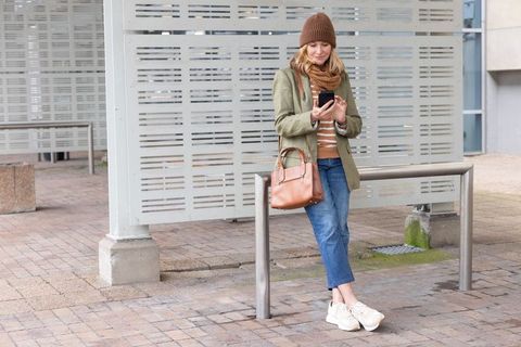 Woman leaning on rail using smartphone in urban plaza wearing olive jacket and tan bag