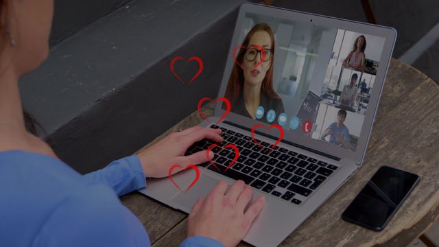 Businesswoman sitting at wooden table, video conferencing on a laptop with multiple colleagues. Red heart icons indicating interactions or relationships are displayed on screen. Useful for illustrating remote work, virtual meetings, technology in the workplace, online communication, and business relationships.