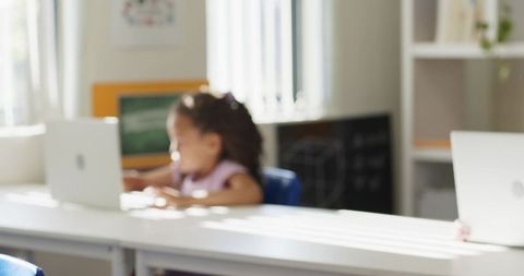 Young girl learning on laptop at sunlit classroom table, focusing on elementary lesson