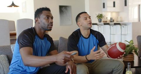 Diverse friends watching football at home on sofa wearing matching blue jerseys and cheering