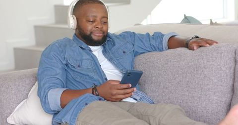 Relaxing african american man listening to music with headphones using smartphone on sofa