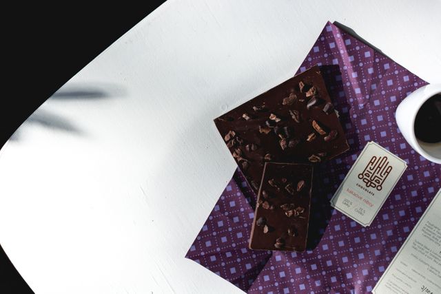 Dark chocolate bar alongside a cup of coffee on a patterned napkin. Ideal for materials promoting gourmet products or illustrating coffee breaks. Great for microblogs about food pairings or elegant table settings. Perfect for use in websites advertising artisanal chocolates or for use in coffee shop promotions.