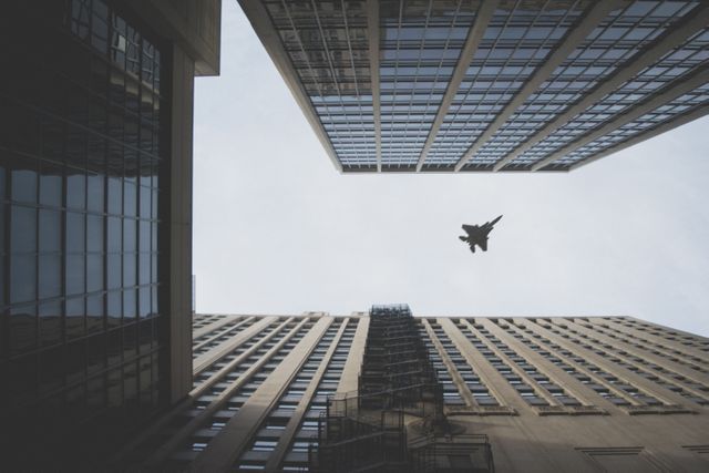 Jet flying through urban canyon between tall skyscrapers with modern glass and concrete structures. Suitable for articles on urban architecture, aviation, and urbanization. Ideal for travel, business, and cityscape projects.