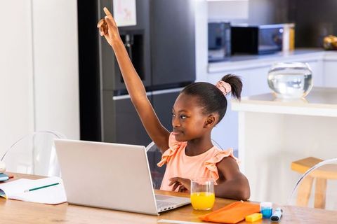 African american girl raising hand studying on laptop in kitchen with glass of orange juice