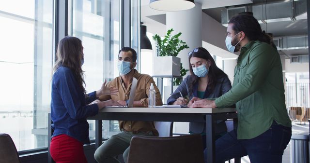 Diverse group of business colleagues wearing face masks sitting at table and talking. working at the office of an independent creative business during covid 19 coronavirus pandemic.
