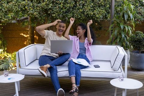 Cheering diverse couple celebrating financial win on patio sofa with laptop and charts