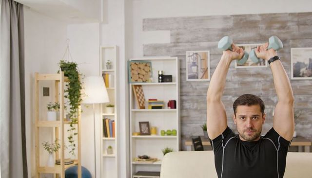 Fit man lifting dumbbells overhead while doing home workout on sofa in cozy living room