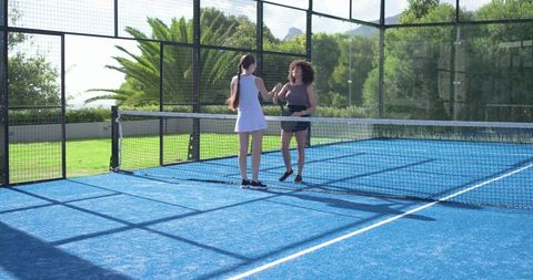 Two diverse female players greeting over net on sunlit blue padel court with glass enclosure