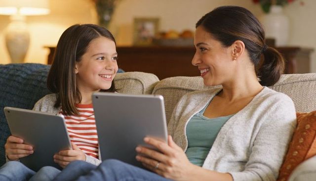 Smiling mother and daughter sharing tablets on couch enjoying cozy family tech bonding moment