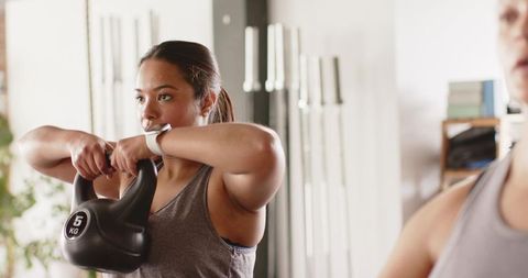 Diverse women lifting 5kg kettlebell in gym studio wearing gray tank tops and smartwatch