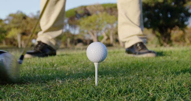 This image shows a close-up perspective of a golf ball on a tee, with a golfer’s feet and golf club in the background, indicating that the golfer is preparing to tee off. This stock photo can be used for advertisements or promotions related to golf, sports equipment, leisure activities, and outdoor events. It is ideal for golf club marketing, sports blogs, and recreational content.