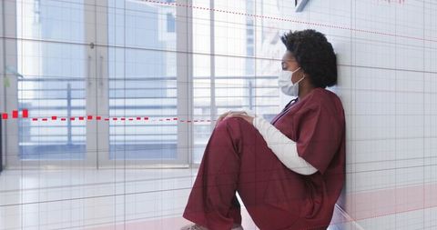 Nurse sitting on hospital floor wearing mask and stethoscope, reflective glass corridor