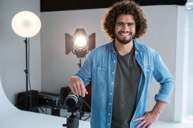 This image depicts a smiling male photographer standing in a studio with professional camera equipment and lighting. Ideal for use in articles or advertisements related to photography, creative professions, studio setups, and professional portraits. It can also be used for promoting photography courses, workshops, or equipment.