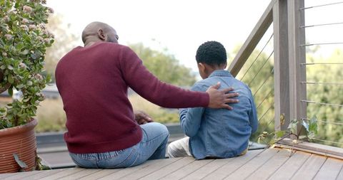 African american father comforting son on wooden balcony deck with greenery and railing