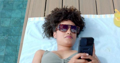 African woman relaxing poolside with sunglasses and smartphone beside citrus drink on sunny deck
