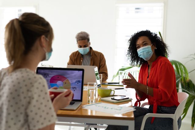 Diverse team members are collaborating in a modern office setting, all wearing face masks for safety. The African American woman is speaking to a Caucasian woman who is holding a cup of coffee, with a laptop displaying a graph in front of her. This image is ideal for illustrating workplace safety during the pandemic, business meetings, teamwork, and professional communication.