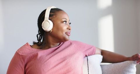 African american woman listening to music on cream headphones while relaxing on pale sofa