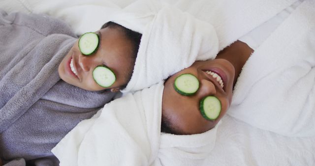 Two women are lying down and relaxing with cucumber slices on their eyes. They are wearing bathrobes and have towels around their heads. This image can be used for spa advertisements, wellness blogs, self-care and beauty publications, or any content promoting relaxation and luxury.