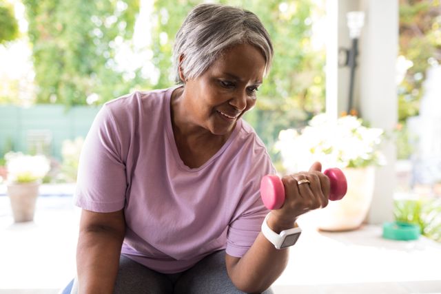 Senior woman lifting a pink dumbbell while sitting in a yard, promoting active lifestyle and healthy aging. Ideal for use in health and wellness articles, fitness blogs, senior care promotions, and advertisements for exercise equipment.
