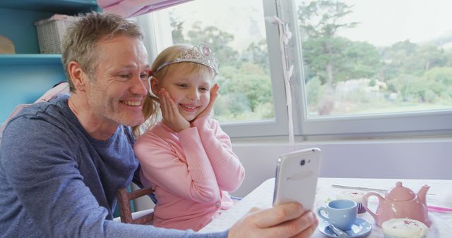 Father and daughter posing for a selfie indoors, with both of them smiling happily. The girl is wearing a pink outfit and a princess crown, indicating a playful and joyous moment. This photo is perfect for illustrating playful family moments, parent-child bonding, marketing family-focused products, or content about happiness and childhood.