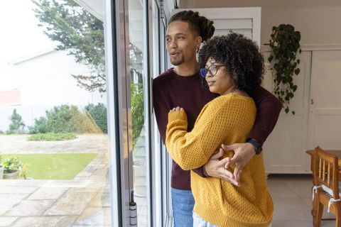 Couple embracing and gazing through sliding door overlooking sunny patio, cozy sweaters