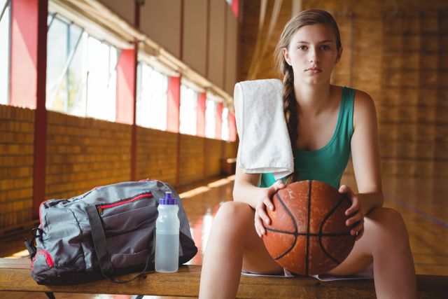 Female basketball player sitting on bench in sports court, holding ball with gym bag and water bottle beside her. Ideal for use in sports-related articles, fitness blogs, athletic training materials, and promotional content for sports equipment or gym facilities.