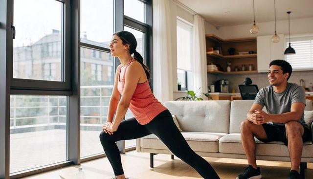 Woman lunging during home workout while partner watches in modern city apartment